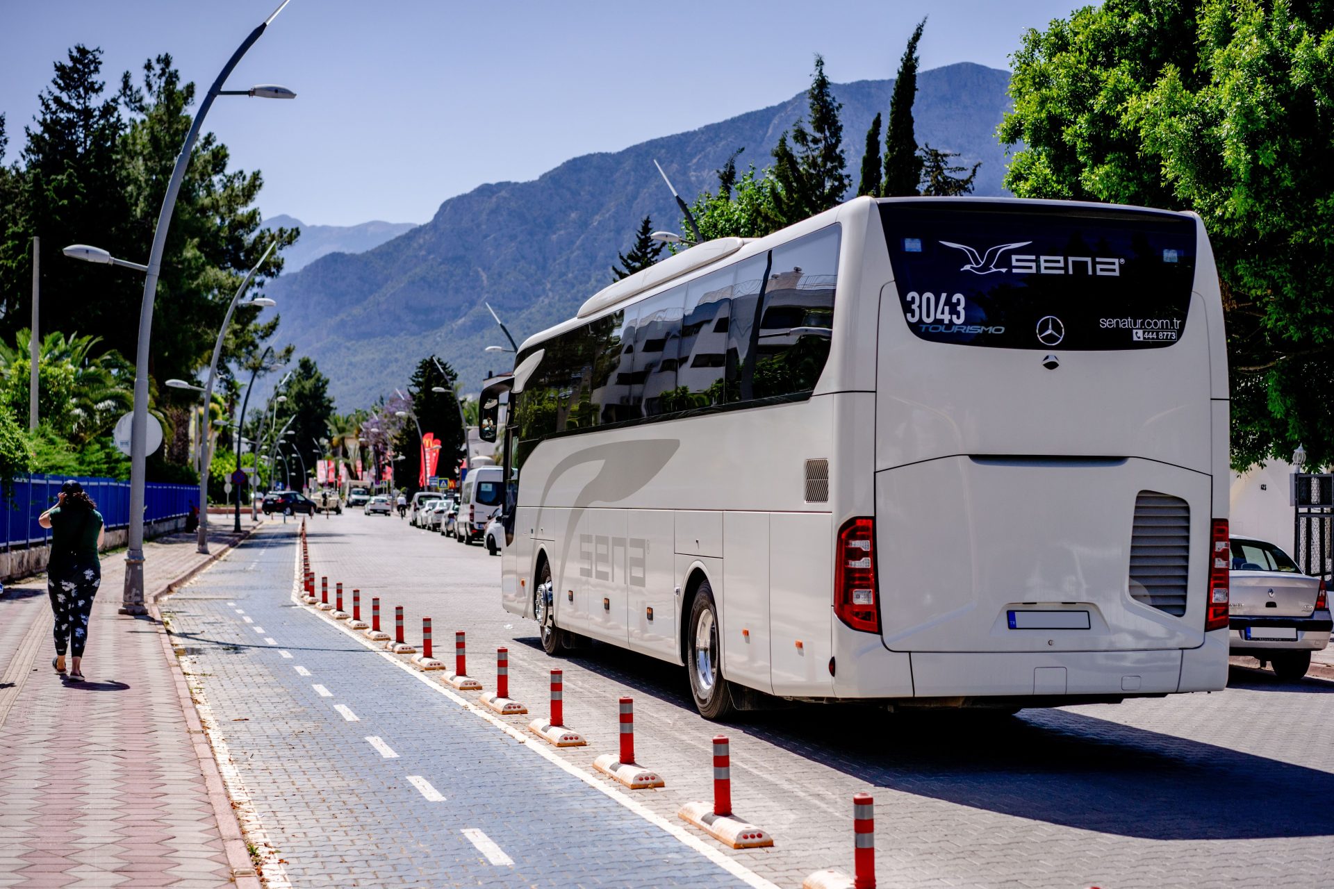 Ein weißer Bus fährt auf einer sonnigen Bergstraße, die von Bäumen und Gehwegen gesäumt ist.